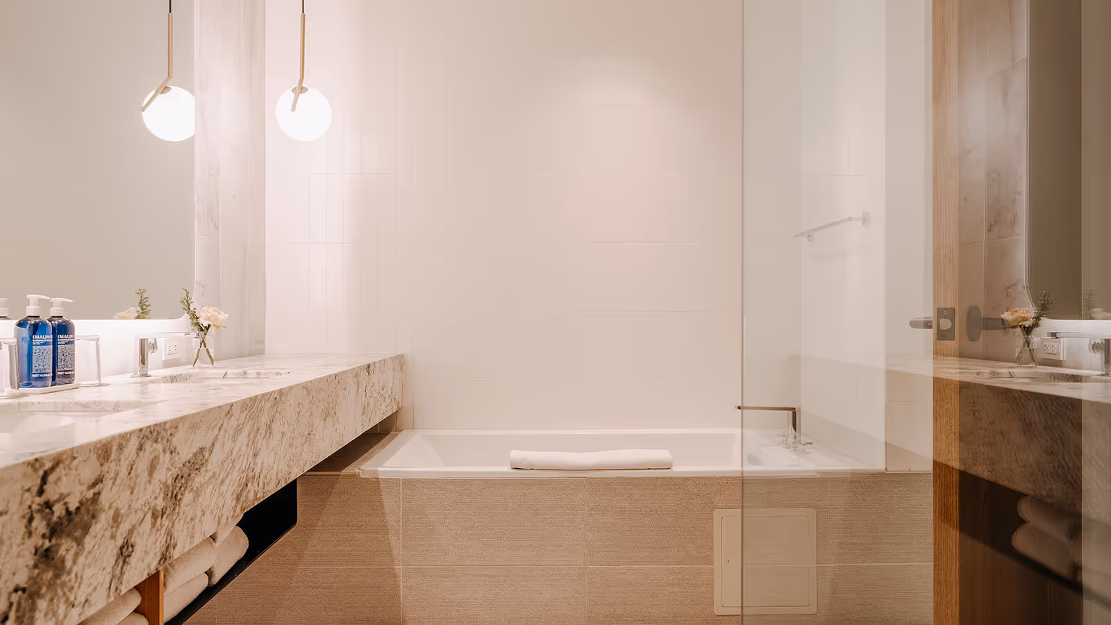 Modern bathroom with marble countertop, built-in bathtub, towel, hanging lights, and toiletries on the counter.