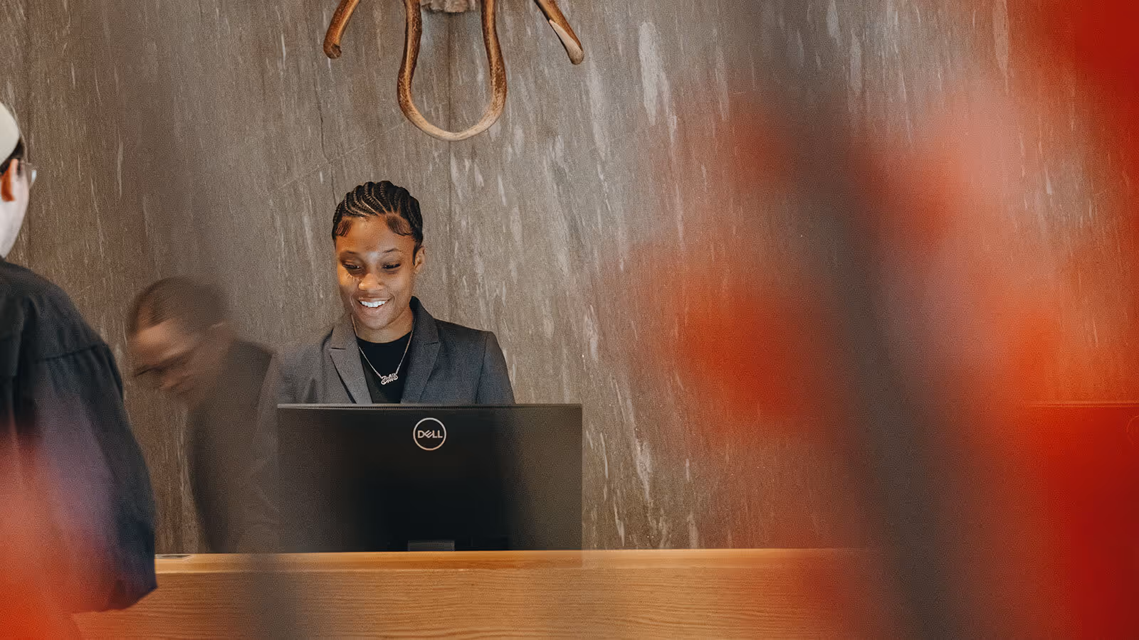 A woman in business attire smiles while working at a reception desk, with a computer and two people nearby.