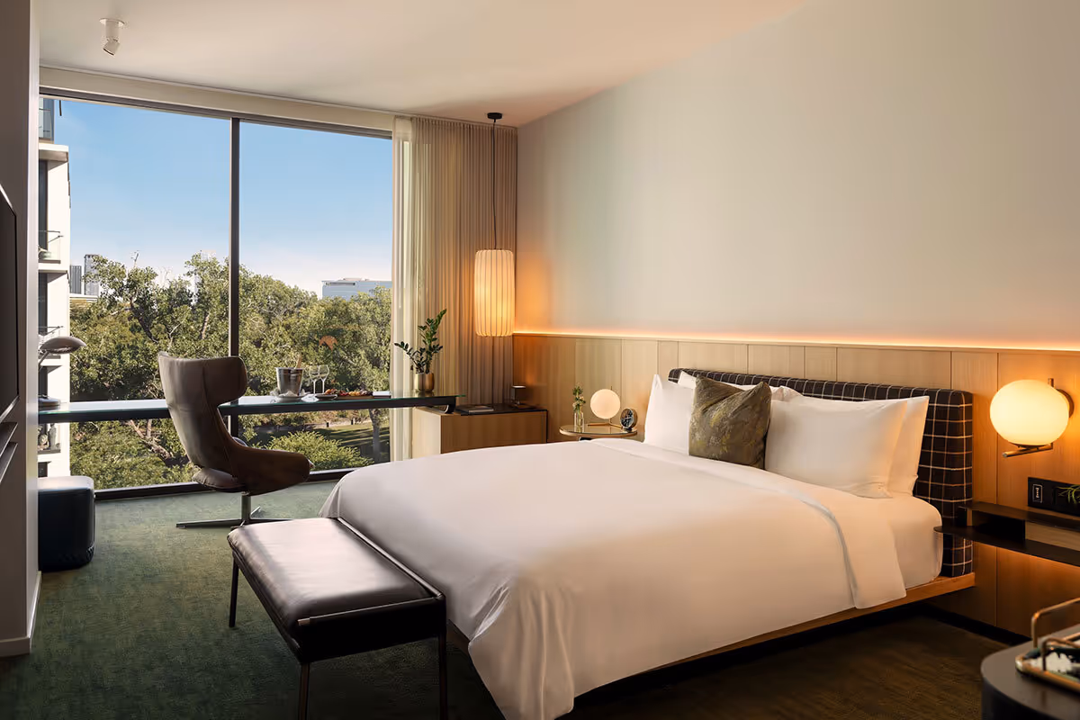 Modern hotel room with a neatly made bed, desk, chair, and large window overlooking trees and buildings.