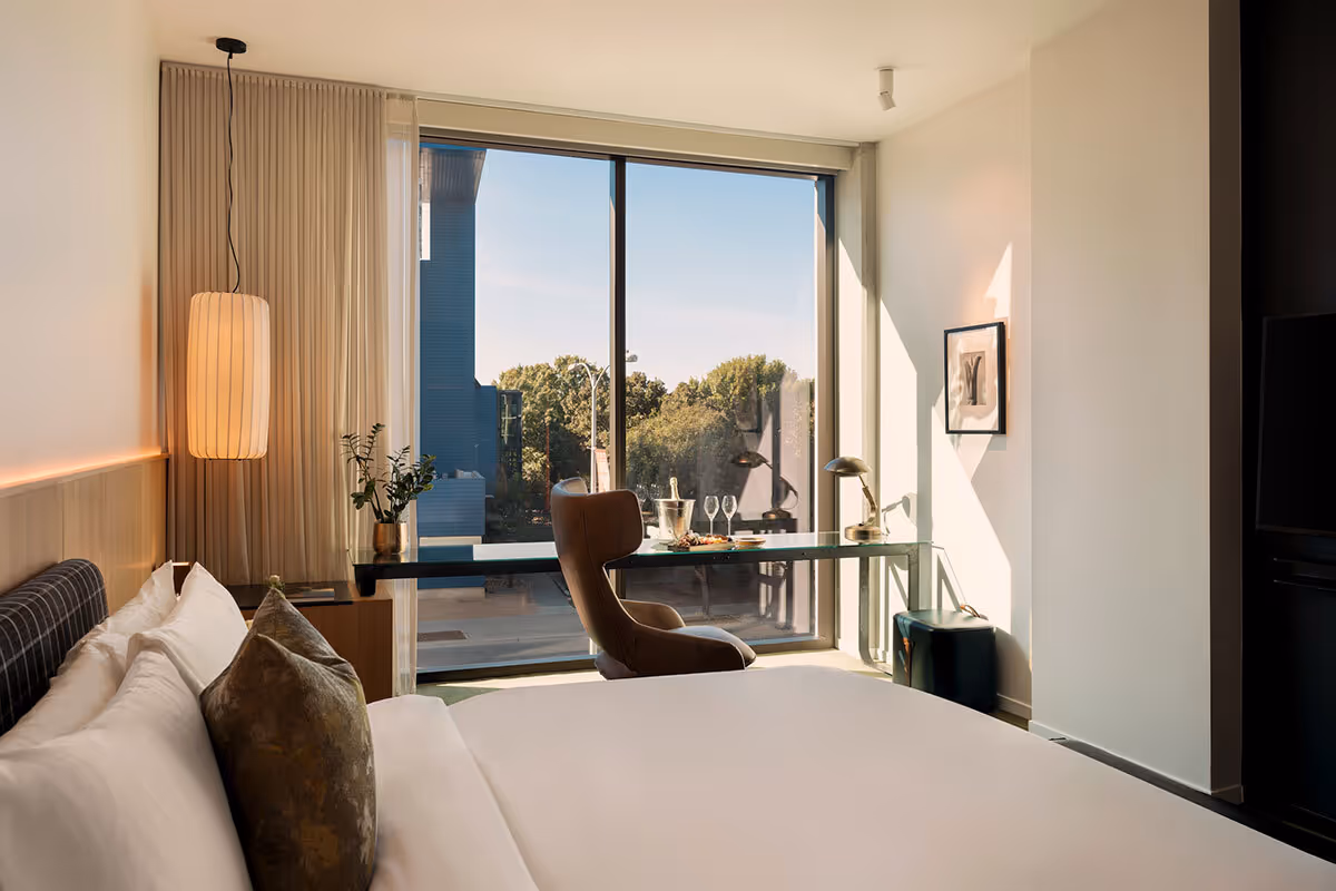 Modern hotel room with a large bed, desk, armchair, and floor-to-ceiling window overlooking trees and a balcony.