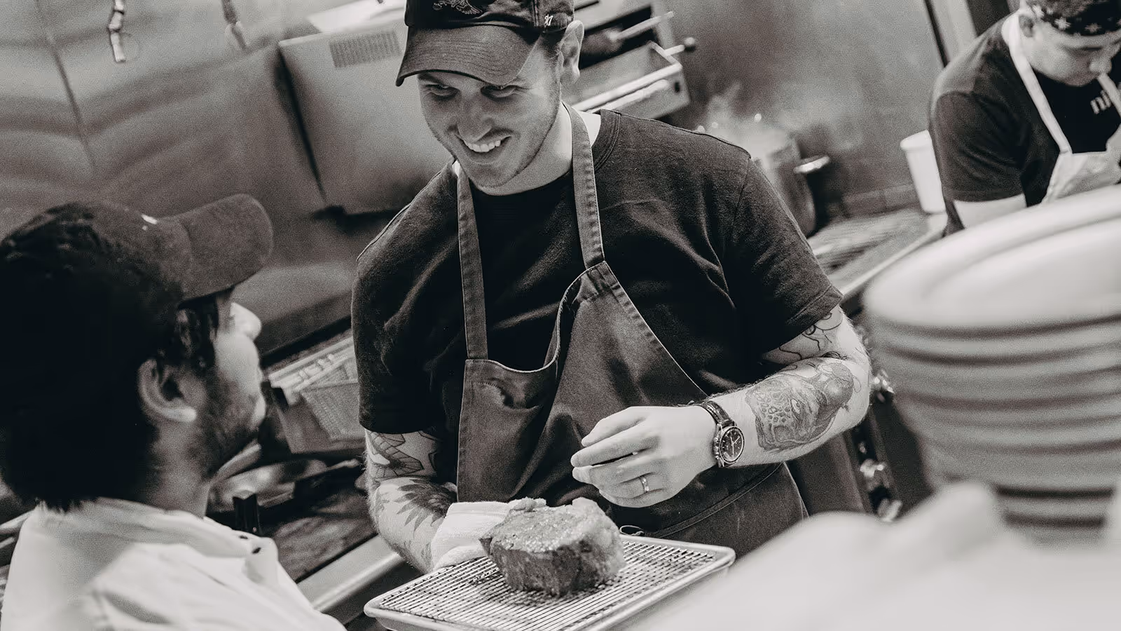 Two chefs in a kitchen talk while one holds a piece of meat on a tray, with plates stacked in the foreground.