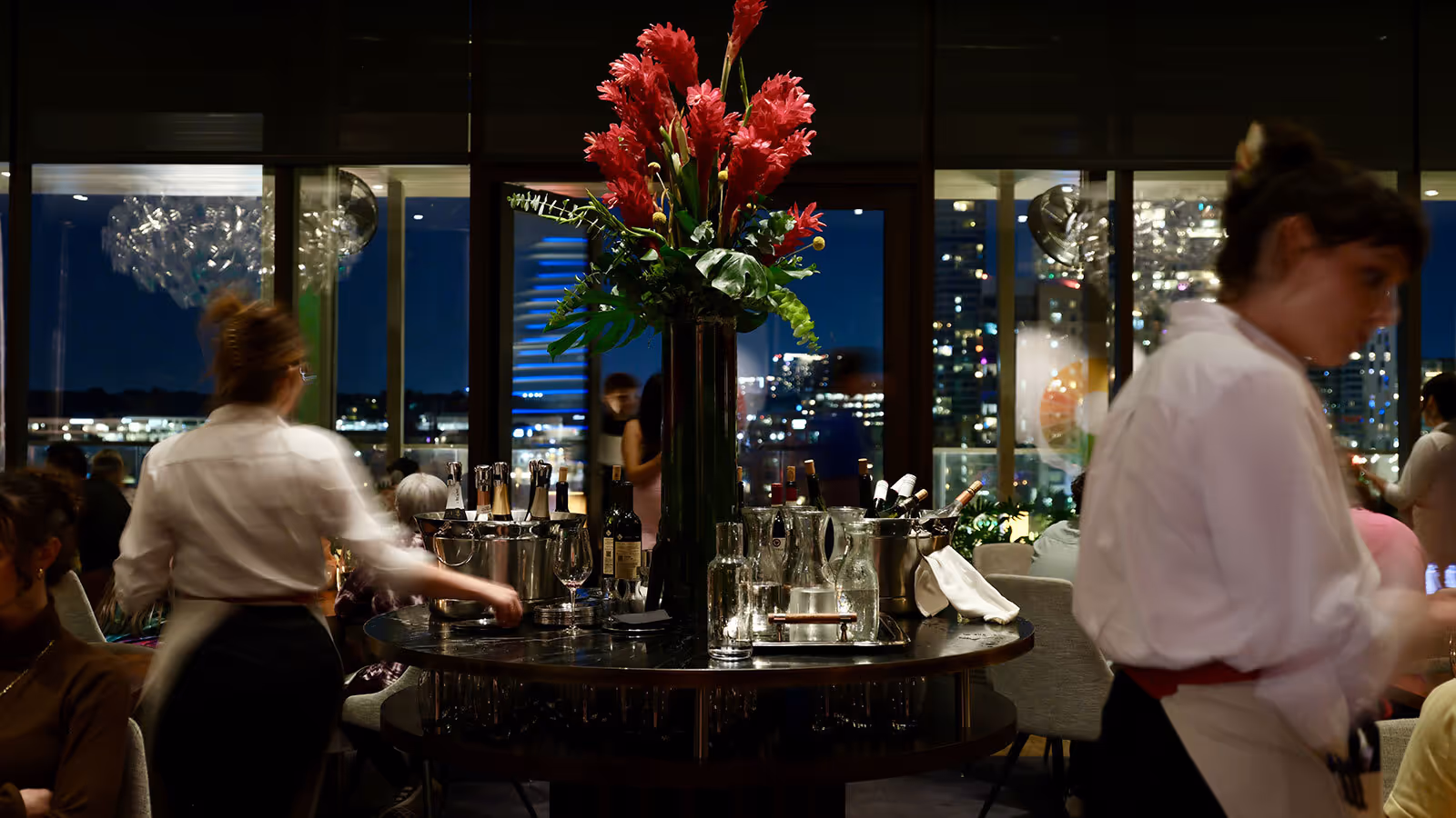 Large flower arrangement on a round table with drinks, surrounded by restaurant staff and diners in a dimly lit setting.