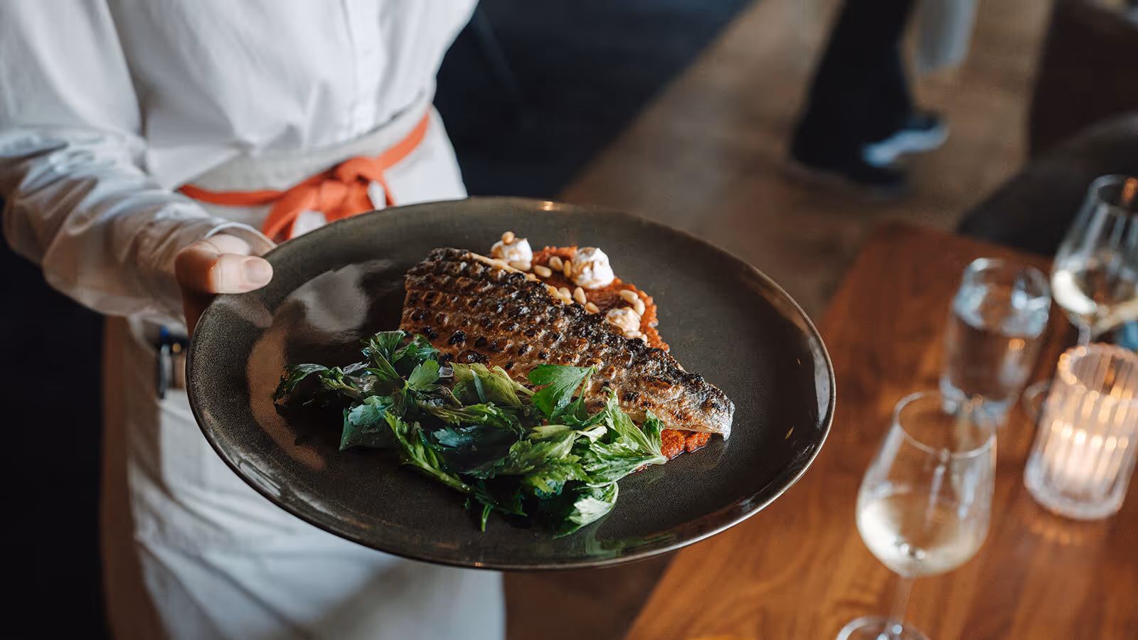 A server carries a plate with grilled fish, fresh greens, and a glass of white wine on a wooden table nearby.