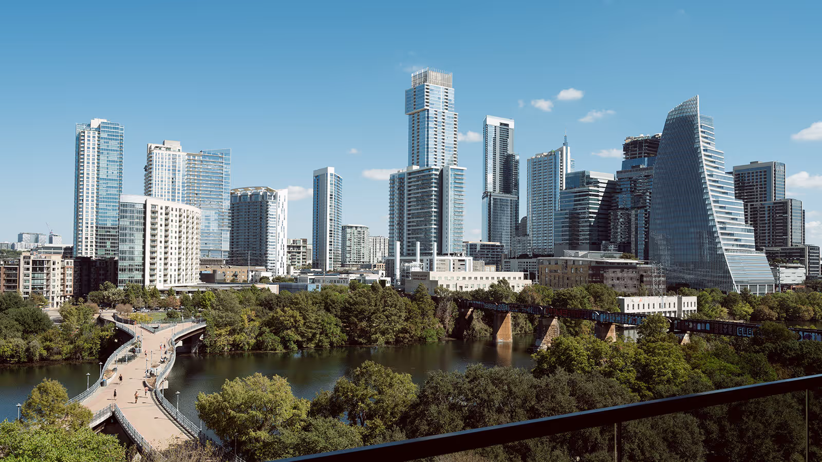View of a modern city skyline with tall buildings, a river, a pedestrian bridge, and trees under a clear blue sky.