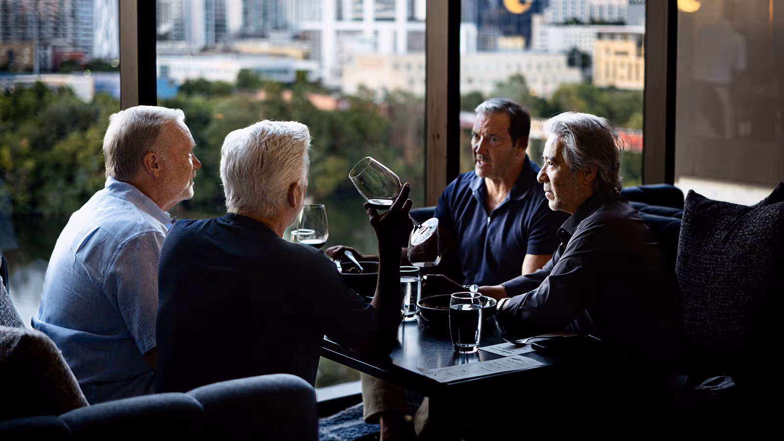 Four men sit at a restaurant table by large windows, talking and drinking wine with a cityscape in the background.