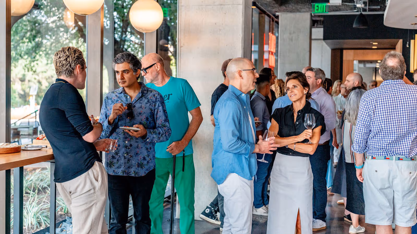 A group of people stand and converse at a social event in a modern indoor space with large windows.
