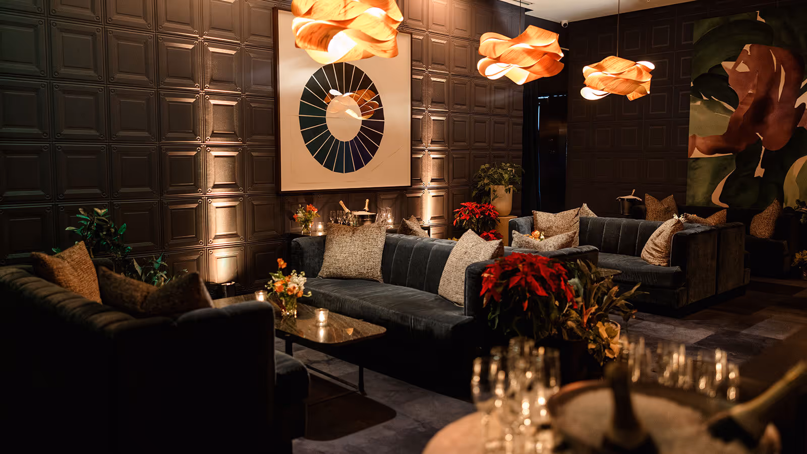 A dimly lit lounge with dark paneled walls, modern sofas, abstract art, and pendant lights above low tables.