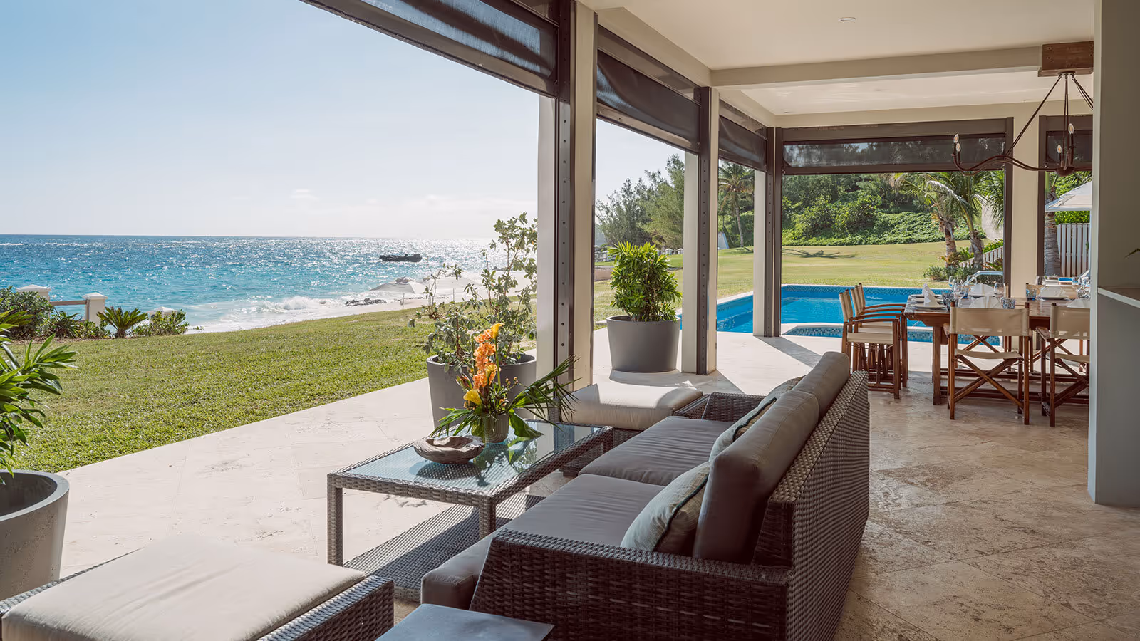Modern patio with wicker furniture overlooks a grassy yard, pool, and ocean view under a clear sky.