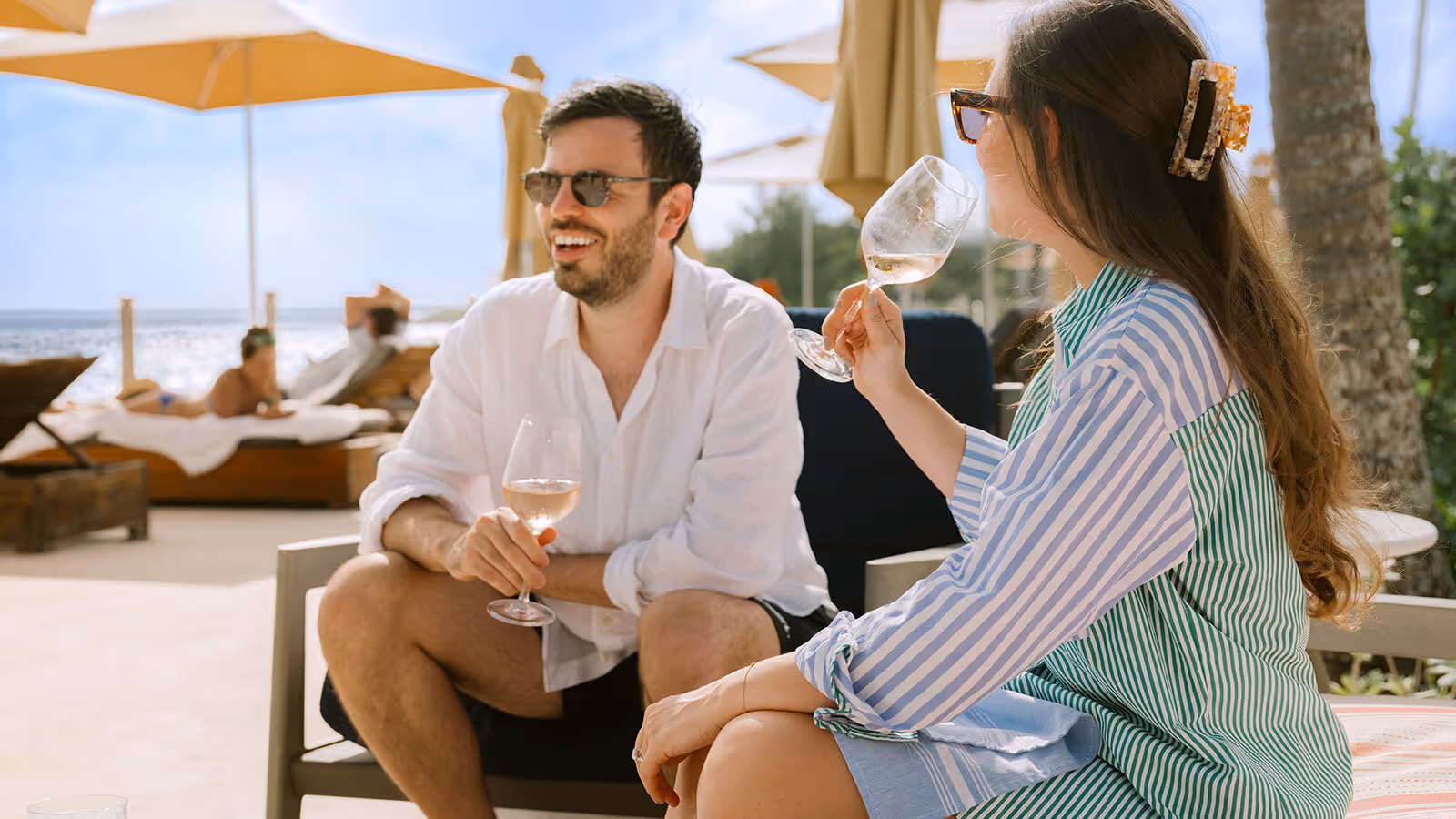 A man and a woman sit outdoors on a beach, relaxing with wine glasses under umbrellas on a sunny day.