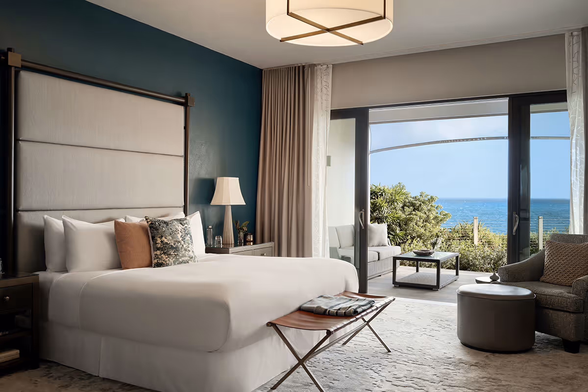 Modern hotel room with a large bed, seating area, and open doors leading to a patio with an ocean view.