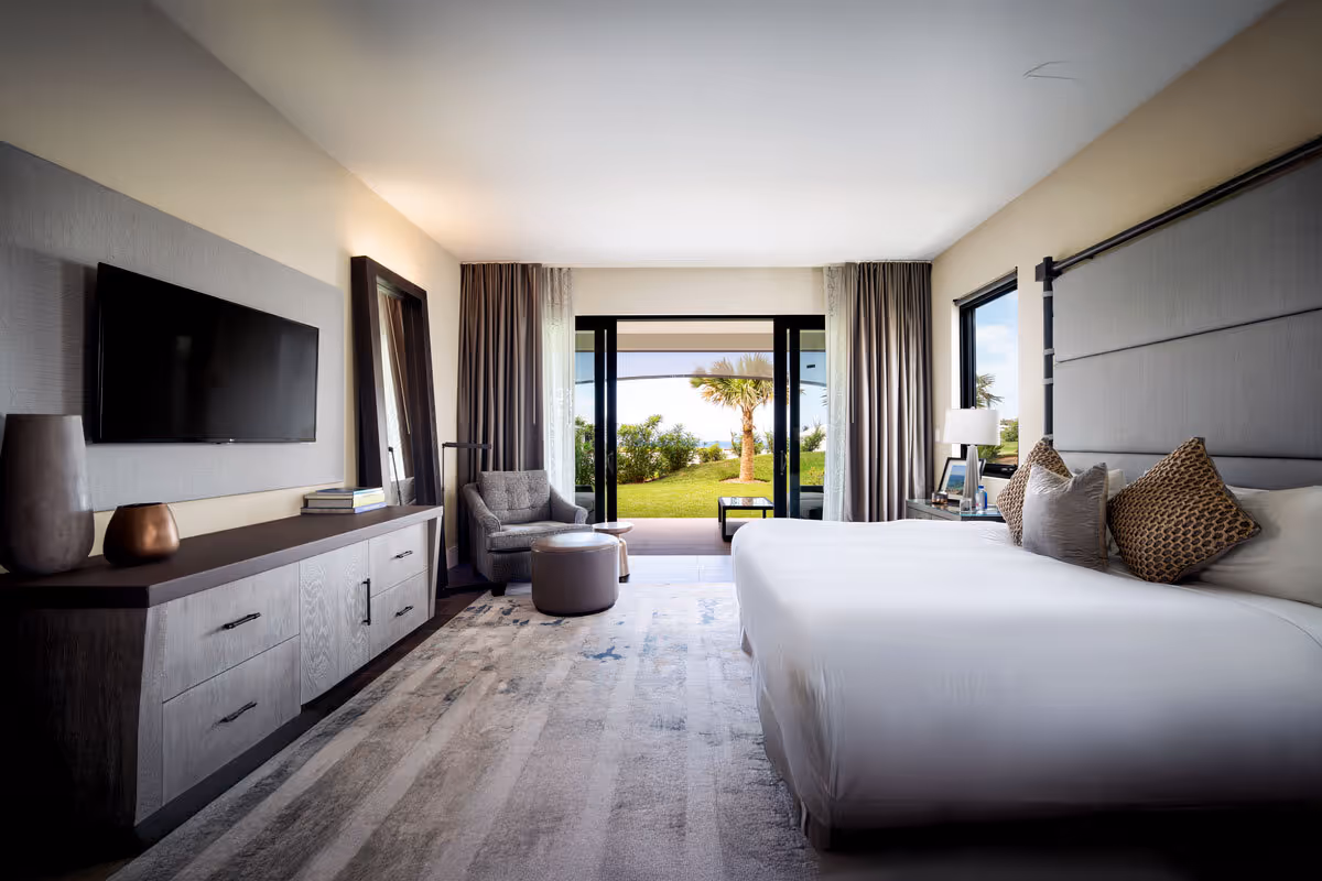 Modern hotel room with a large bed, flat-screen TV, lounge chair, and balcony view of greenery and palm trees.