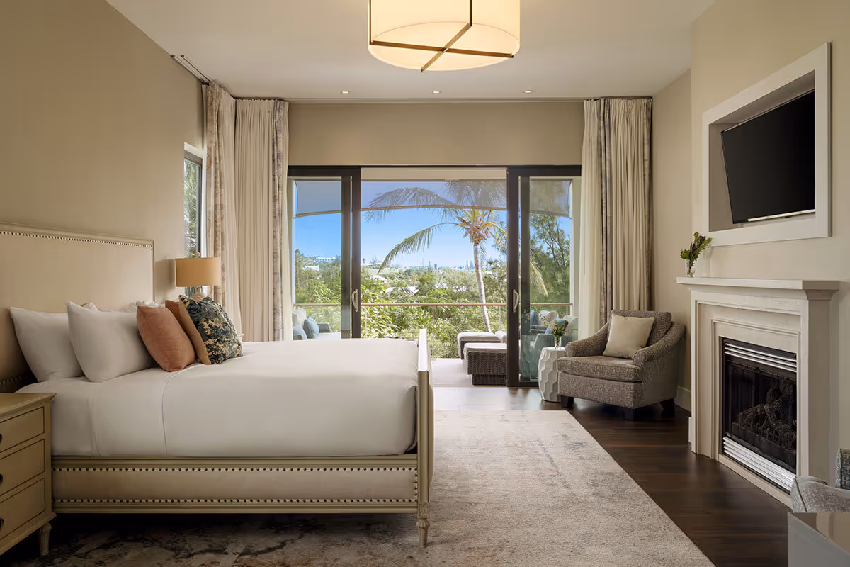 Modern bedroom with large bed, armchair, fireplace, wall-mounted TV, and glass doors opening to a balcony with a palm view.