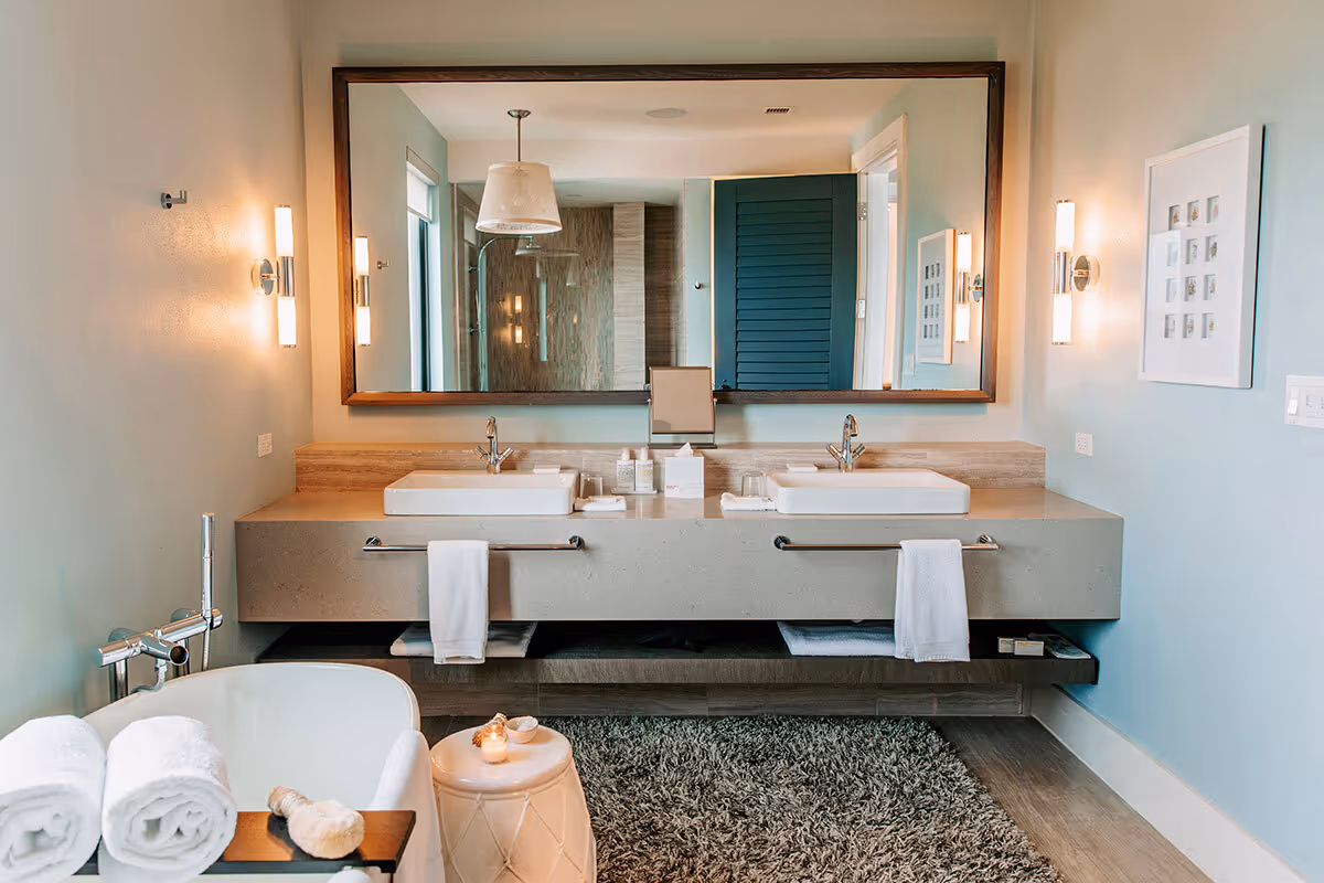 Modern bathroom with double sinks, large mirror, bathtub, wall lights, and towels on a rug-covered floor.