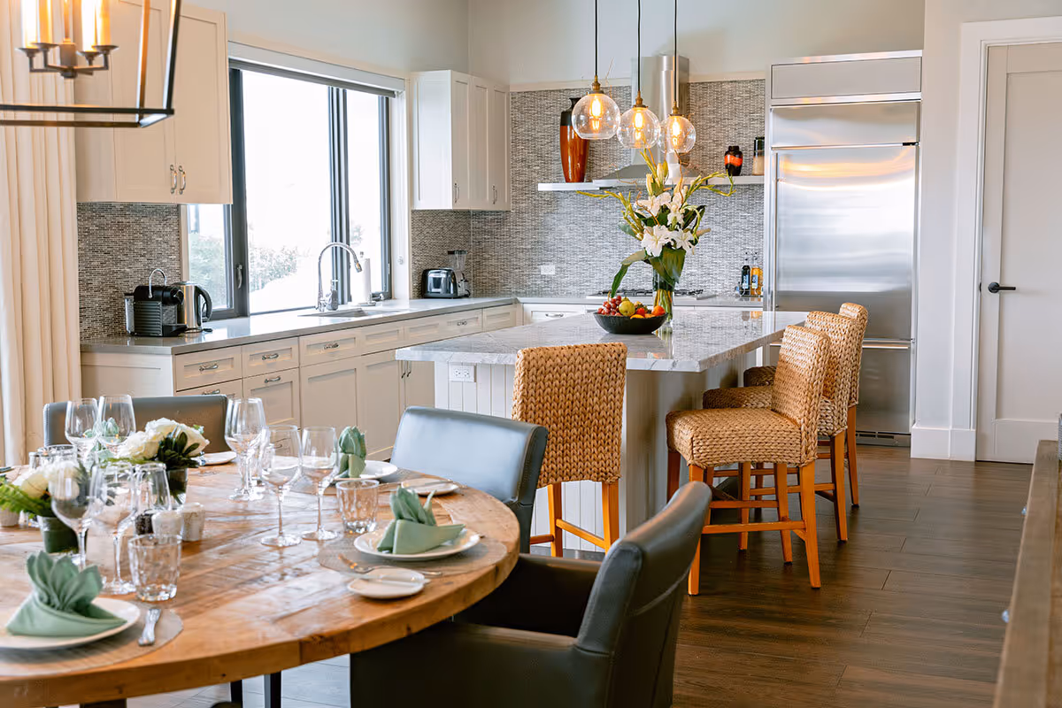 Modern kitchen with island, woven barstools, stainless appliances, and a set dining table in the foreground.