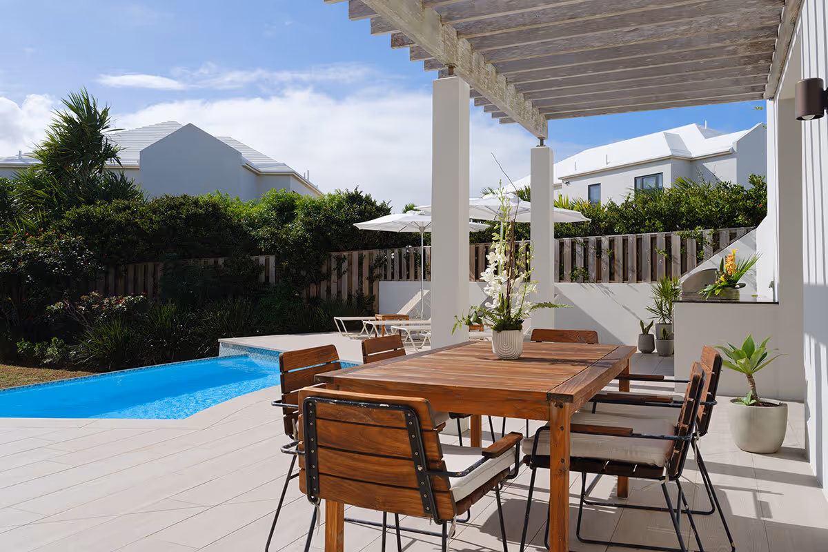 A wooden outdoor dining table and chairs on a patio beside a swimming pool, with houses and greenery in the background.