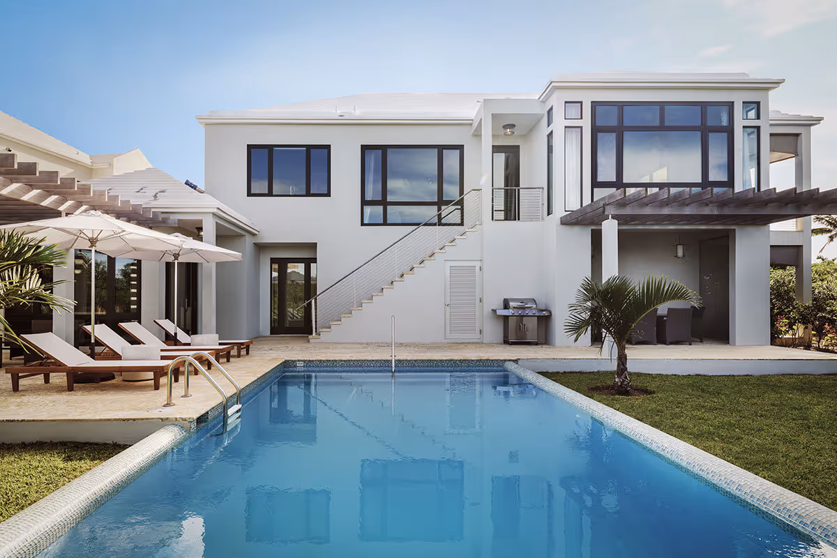 Modern two-story house with large windows, a swimming pool, lounge chairs, and a small palm tree in the backyard.