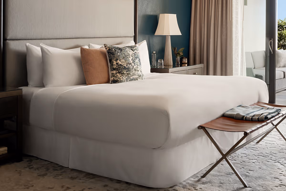A neatly made bed with white linens, decorative pillows, a bedside lamp, and a bench at the foot of the bed.