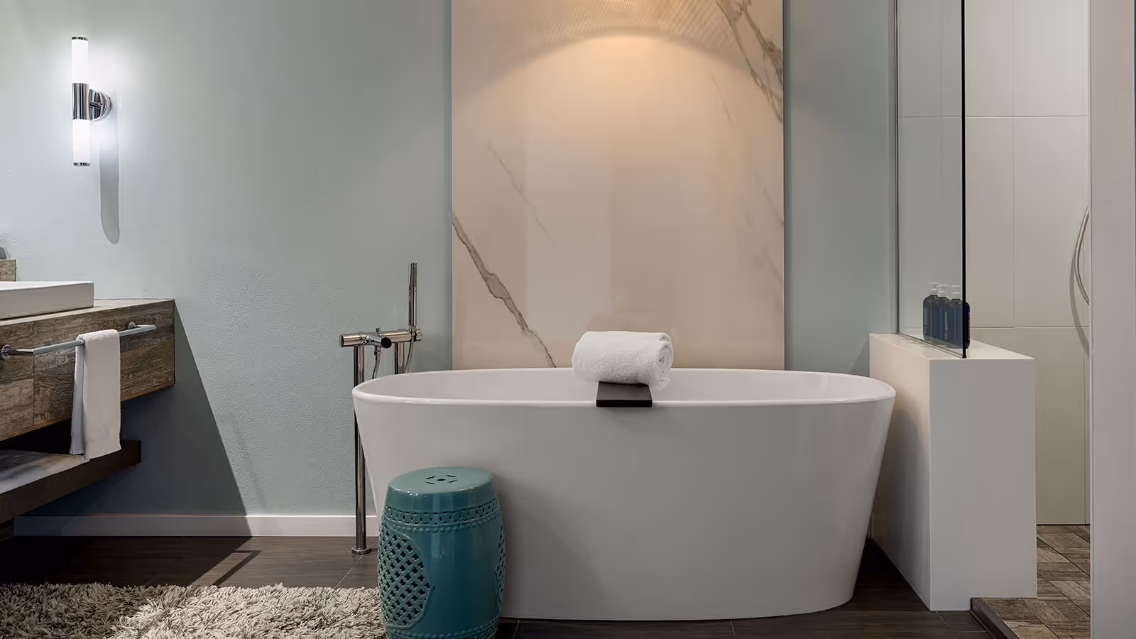 Modern bathroom with a freestanding white tub, towel, green stool, wall light, and textured rug on dark wood floor.