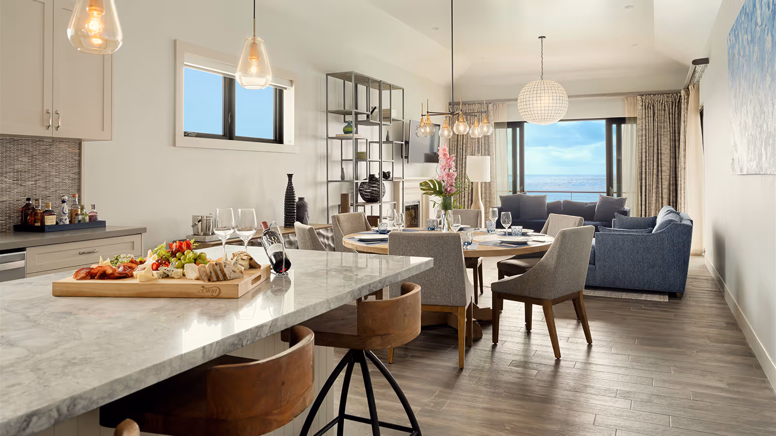 Modern open-concept kitchen and dining area with marble island, wooden chairs, and ocean view through large windows.