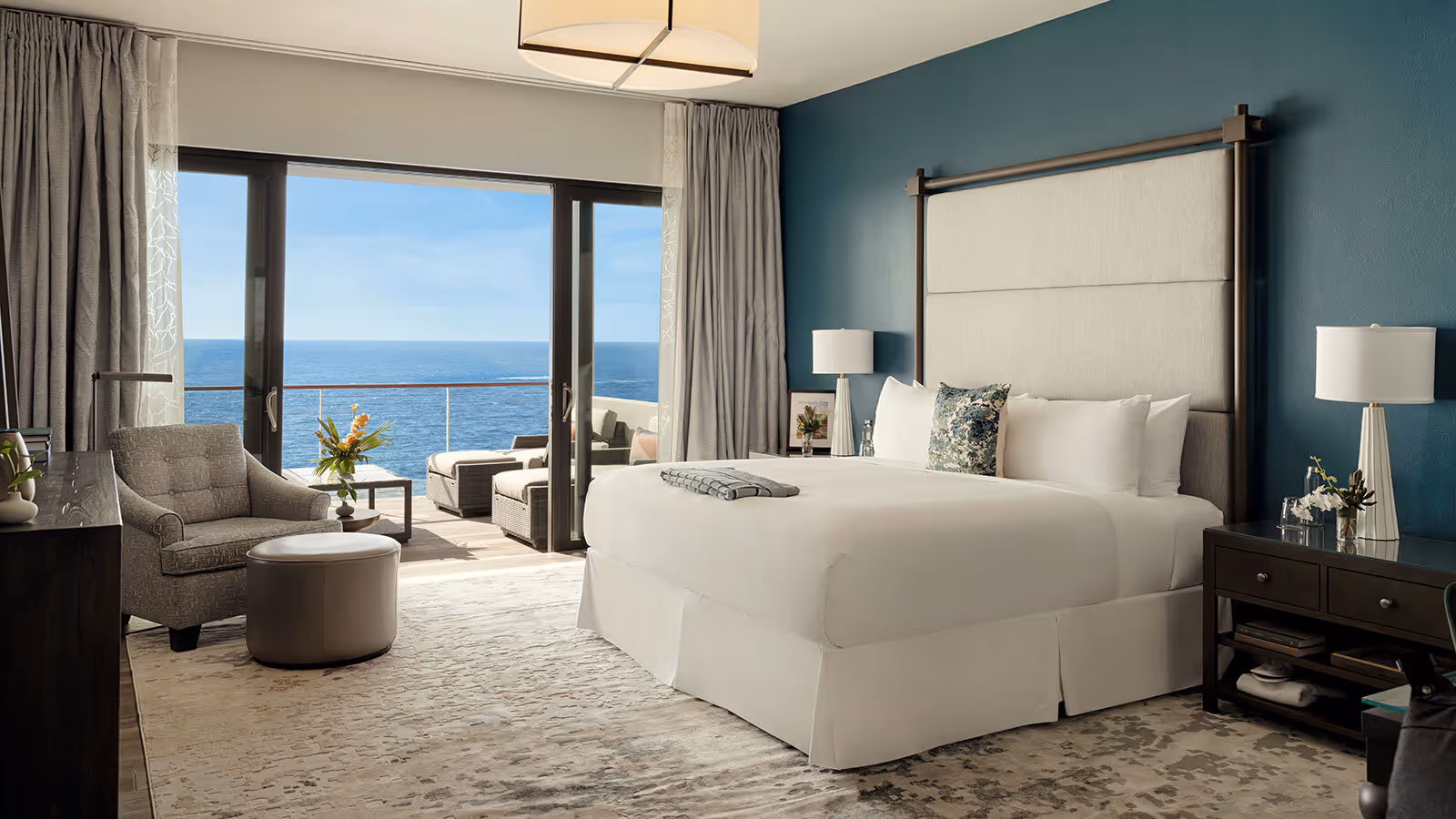 Modern hotel room with a large bed, armchair, and balcony overlooking the ocean under a clear blue sky.