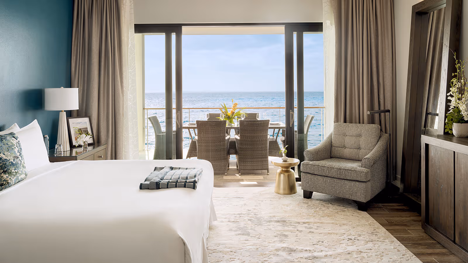 A hotel room with a bed, armchair, and balcony overlooking the ocean with a dining table and chairs outside.