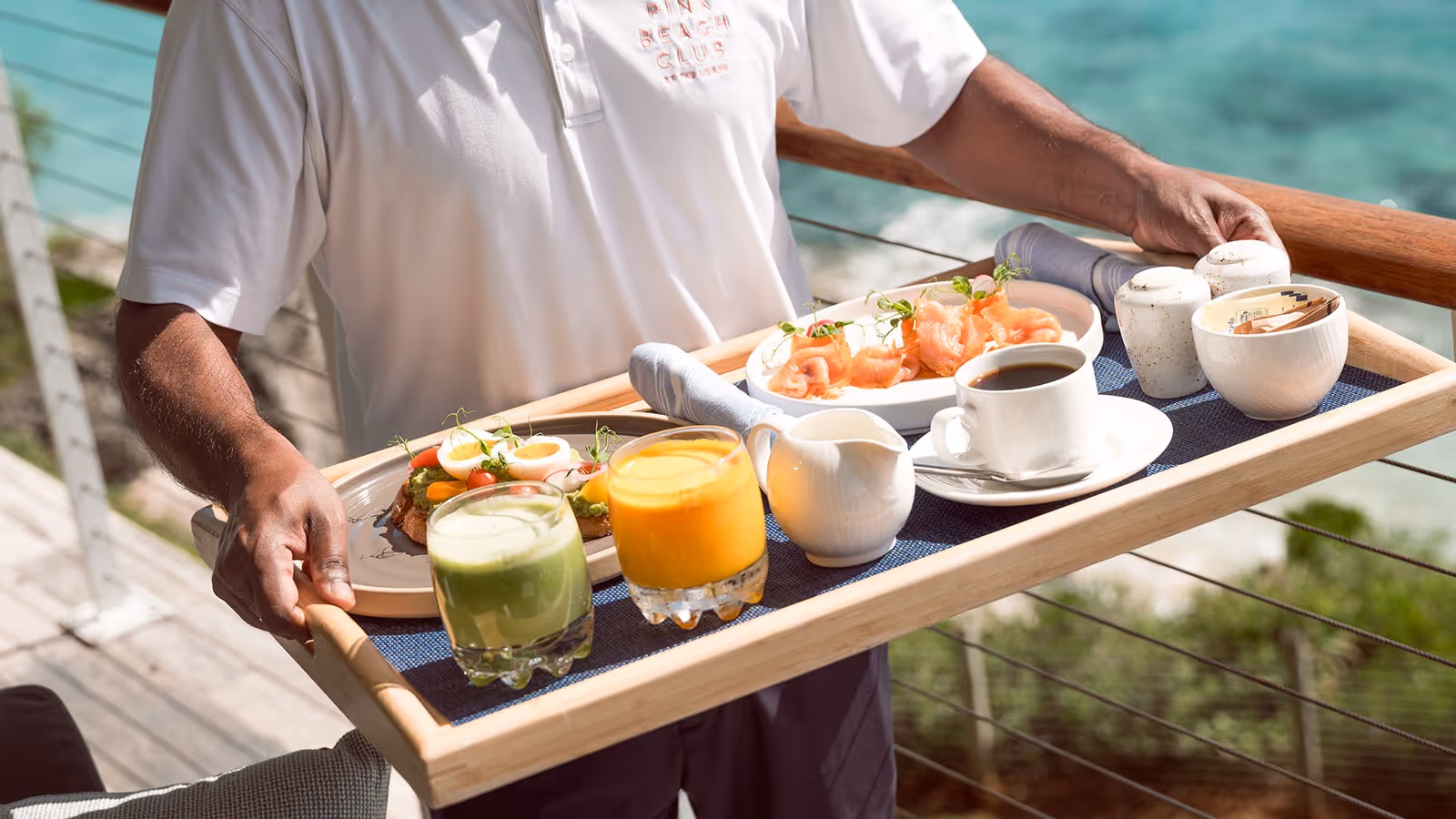 Person holding a tray with juice, coffee, yogurt, smoked salmon, toast, and napkins on an outdoor balcony by the ocean.