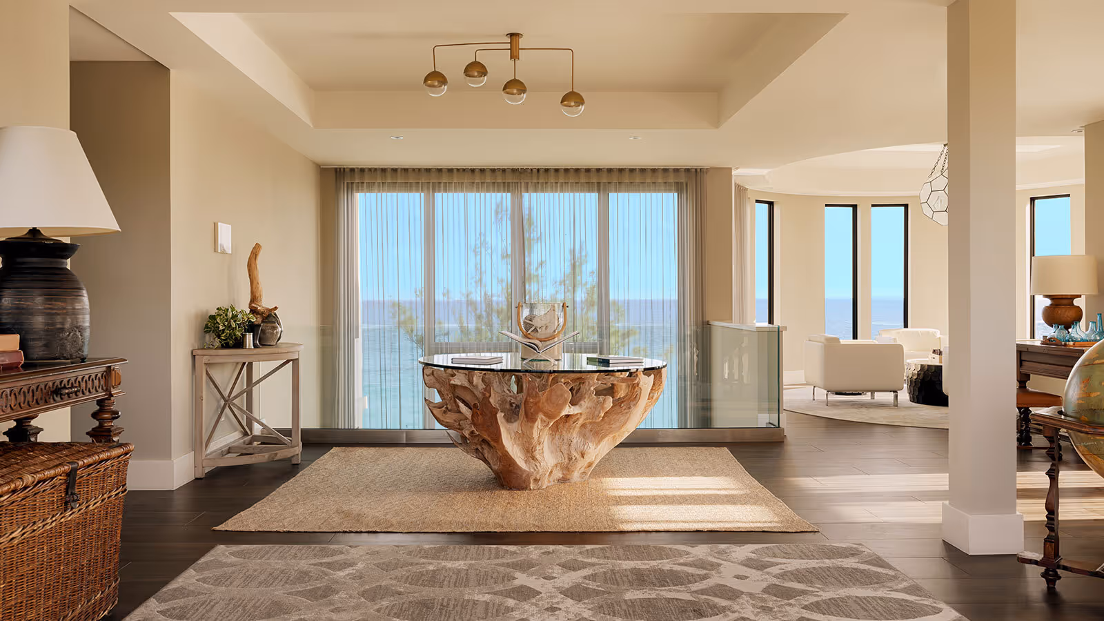 Modern living room with a large driftwood base glass table, coastal decor, and floor-to-ceiling windows overlooking the ocean.