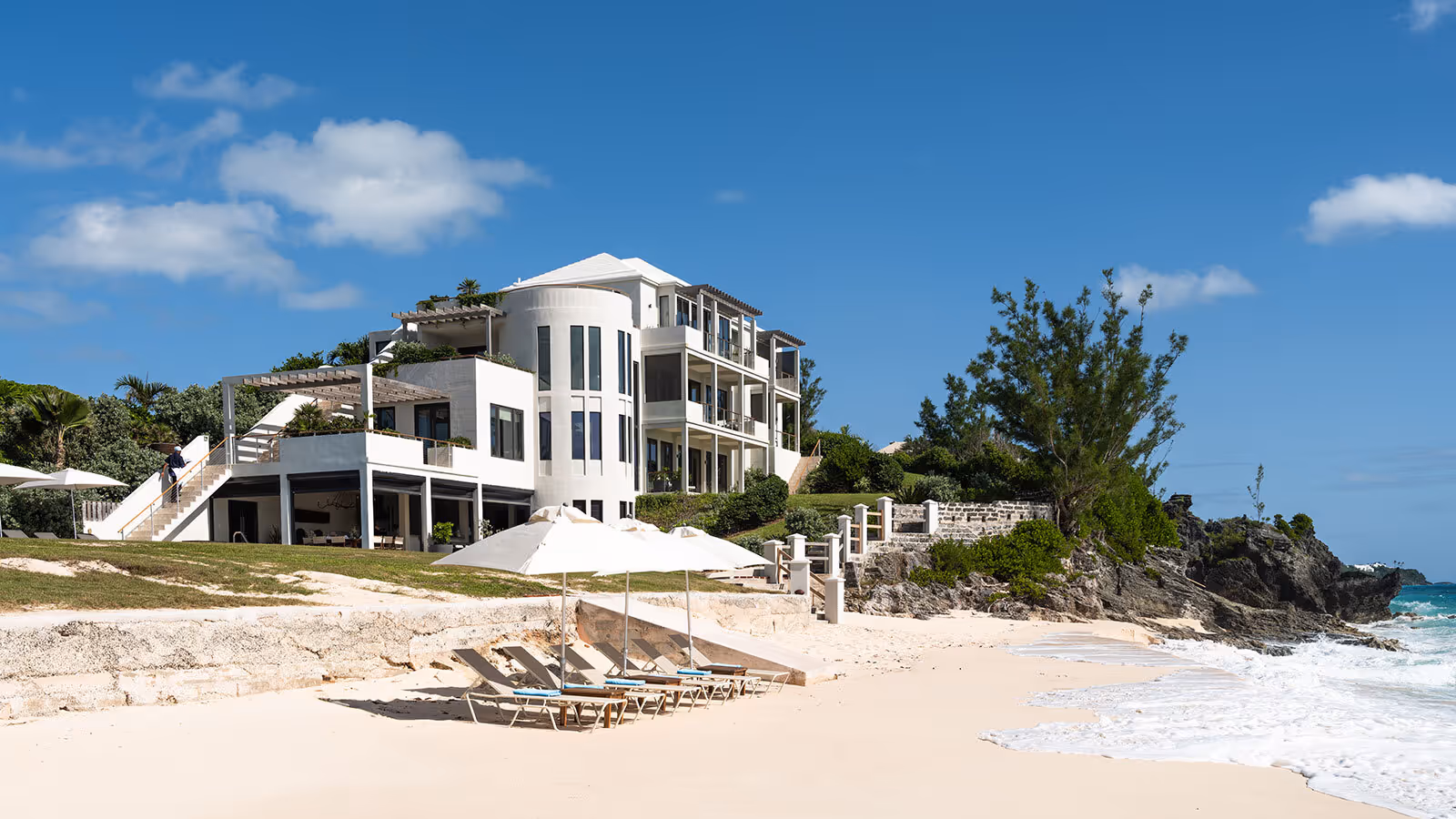 Modern white beachfront villa with multiple balconies, large windows, and lounge chairs under umbrellas on a sandy beach.