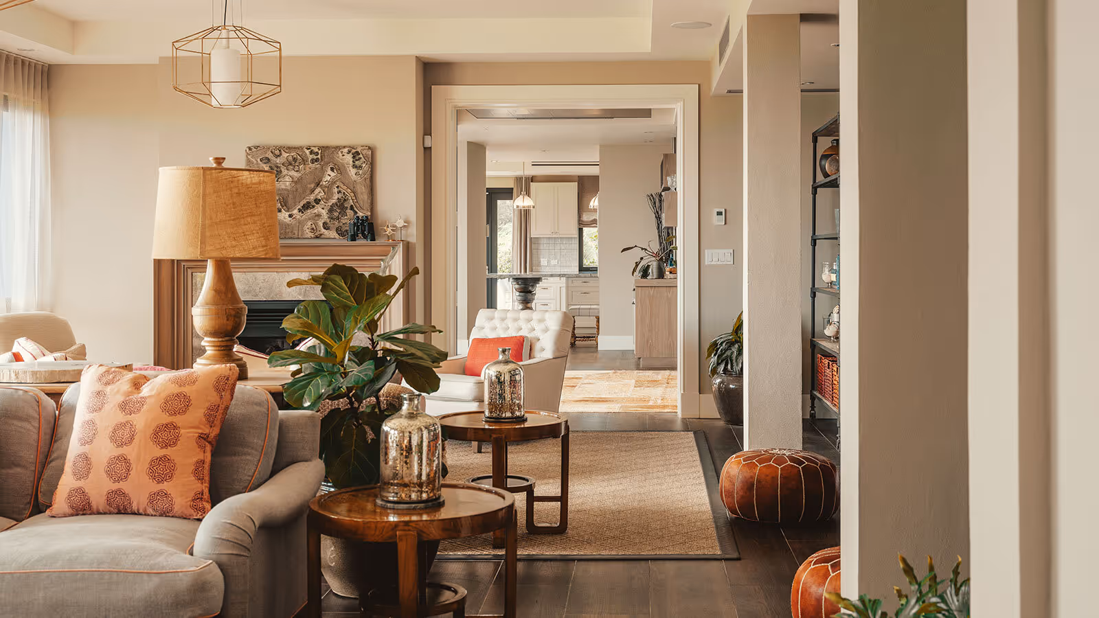 A modern living room with neutral tones, plants, patterned pillows, and a view into a bright kitchen.