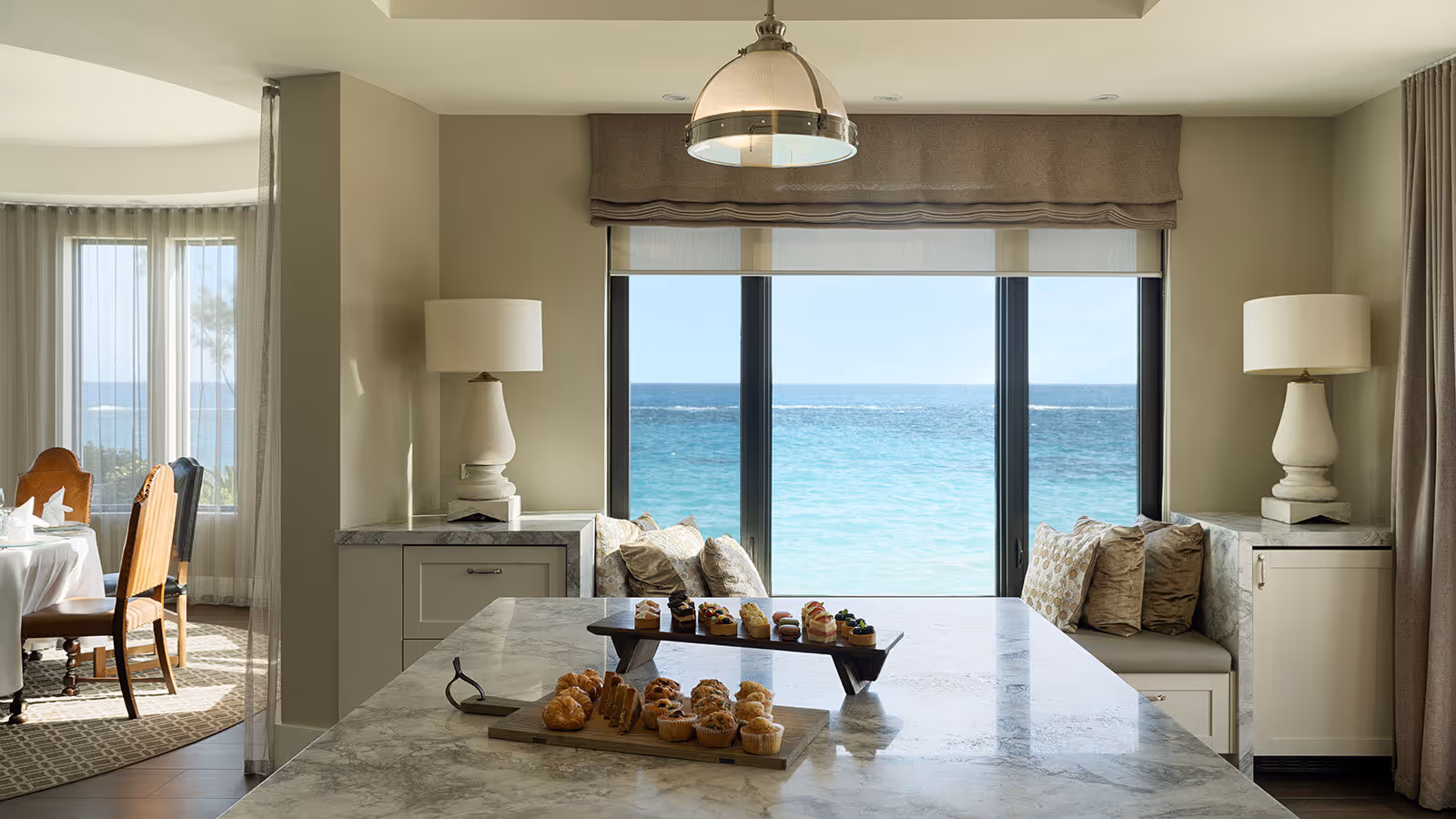 Marble kitchen island with pastries, large windows overlooking the ocean, and window bench seating with pillows.