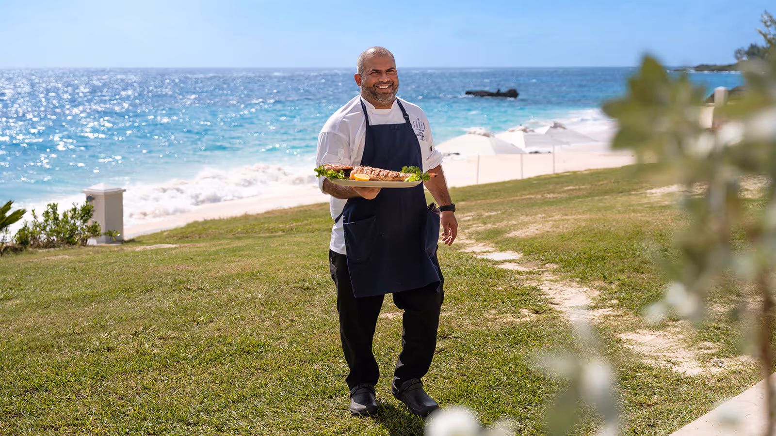 A chef in uniform stands on grass by the ocean, holding a platter of food and smiling at the camera.