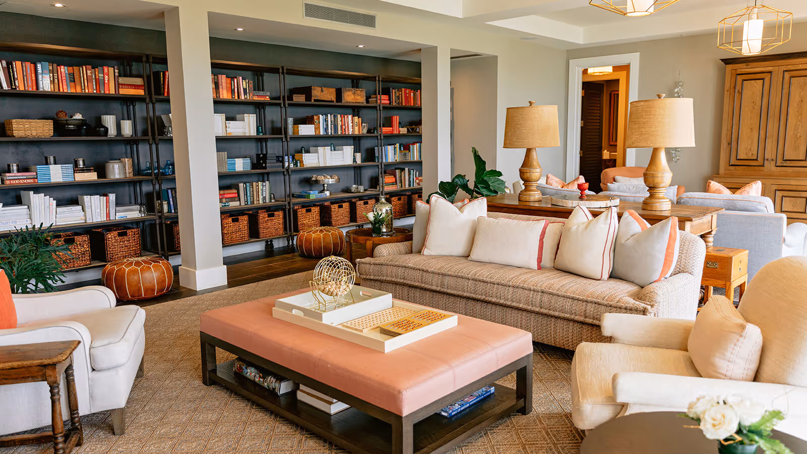 A cozy living room with neutral-toned sofas, a pink ottoman, and large bookshelves filled with books and baskets.