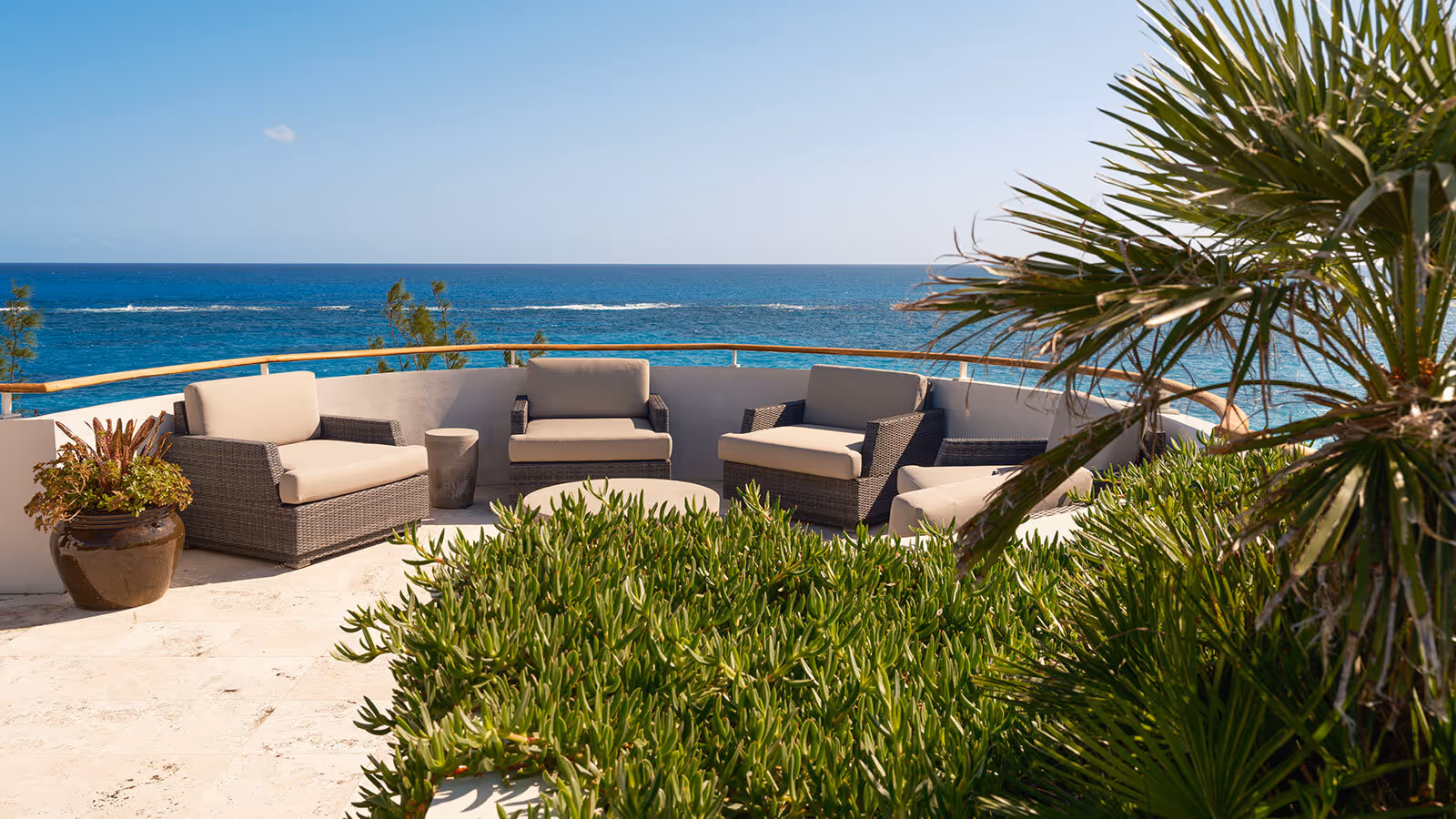 Outdoor patio with wicker chairs and sofas overlooks the ocean, surrounded by green plants under a clear blue sky.
