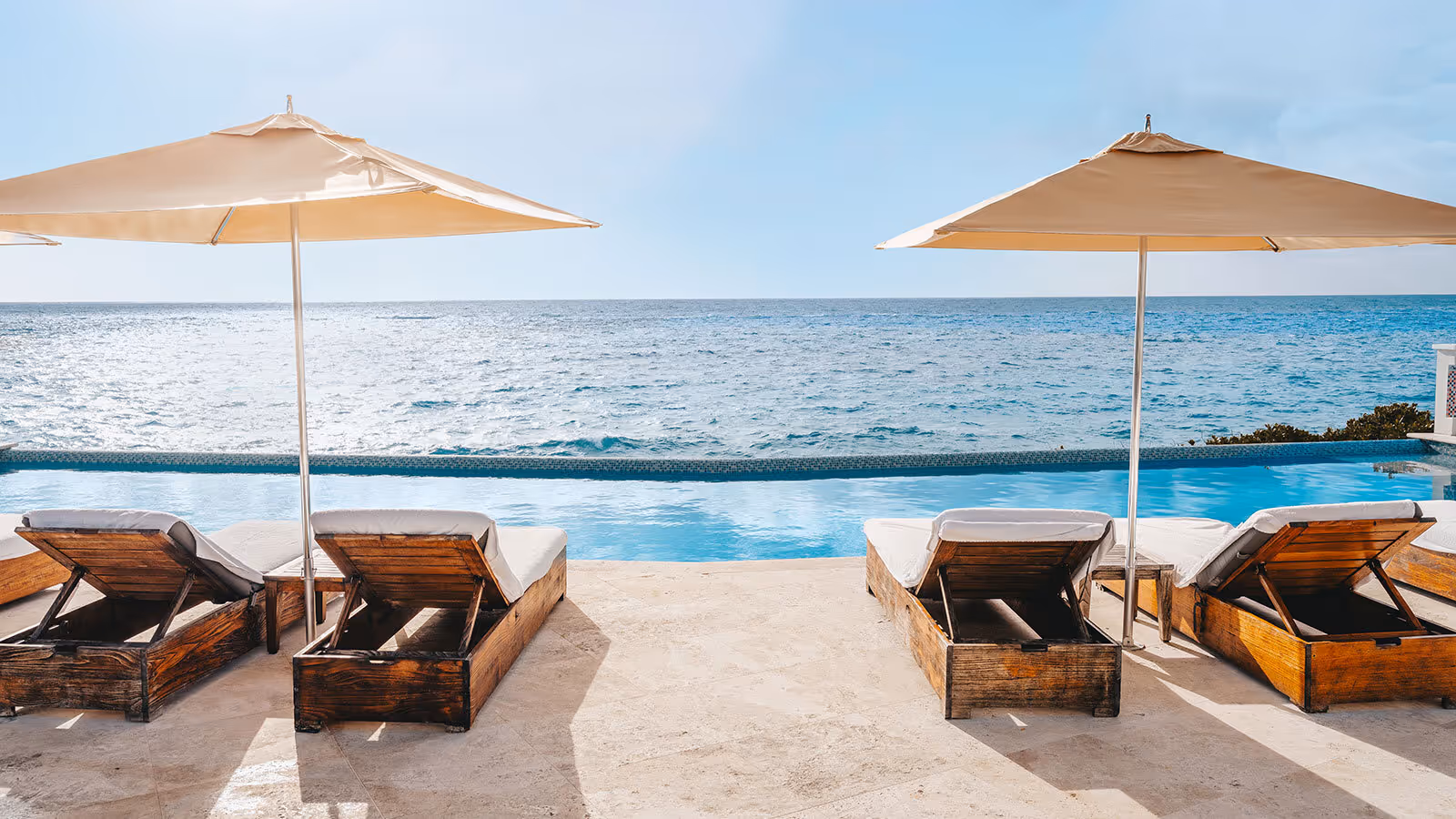 Two wooden lounge chairs under umbrellas face an infinity pool and a calm ocean on a sunny day.