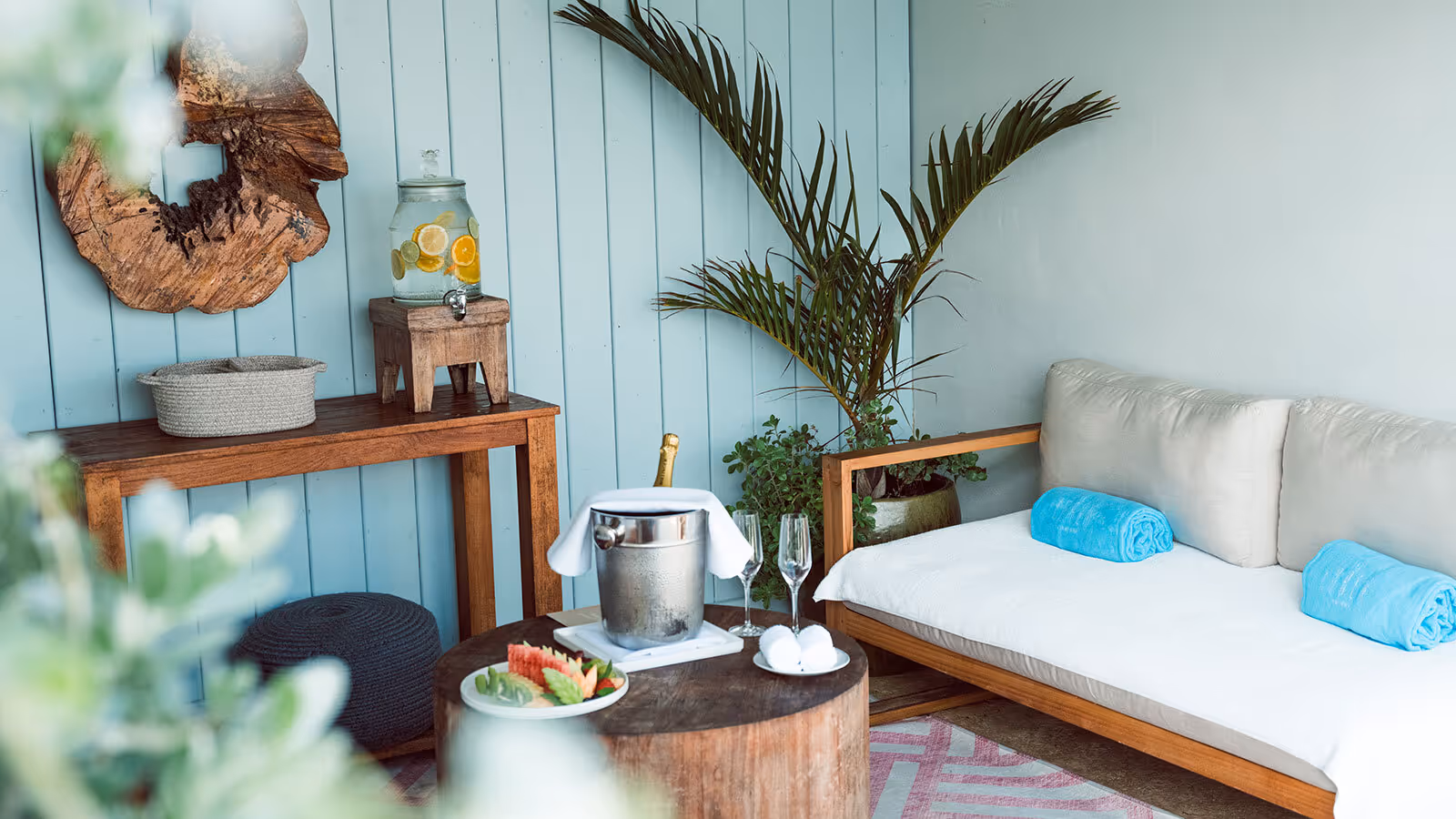 Outdoor seating area with a white cushioned sofa, wooden table, ice bucket, two glasses, and a water dispenser.