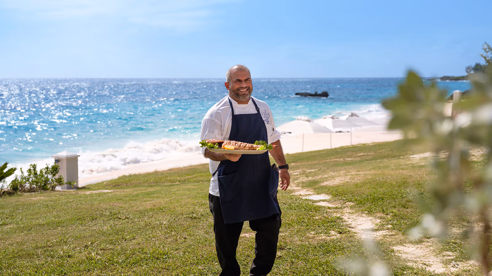 A chef in uniform stands on grass by a beach, holding a platter of food, with the ocean in the background.