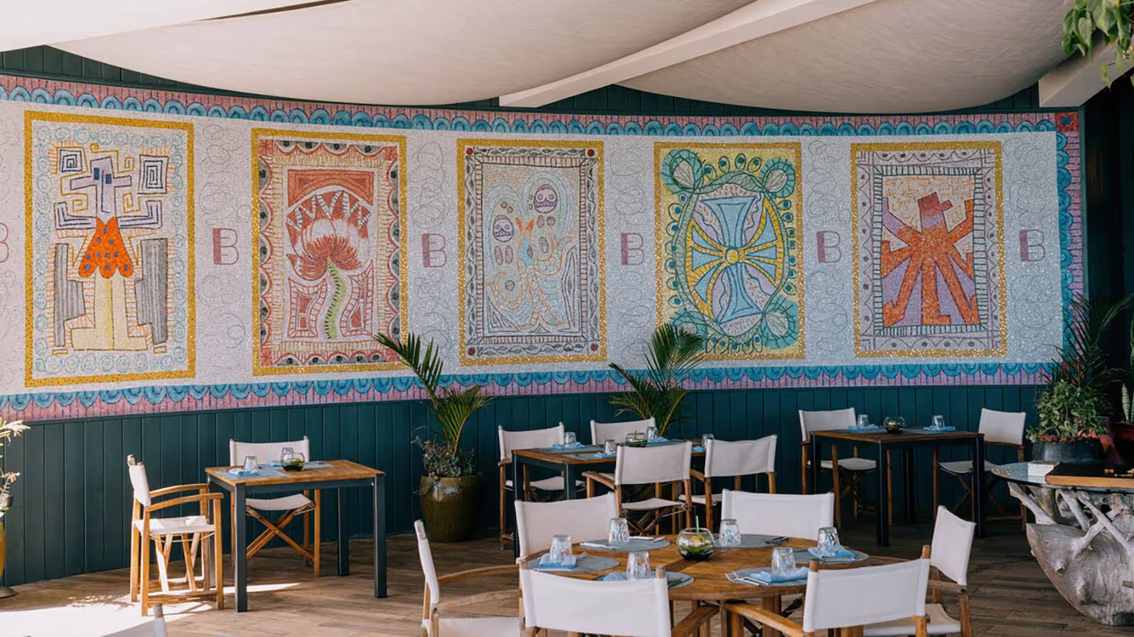 Outdoor dining area with wooden tables and white chairs, featuring a colorful mural with five large, patterned panels on the wall.