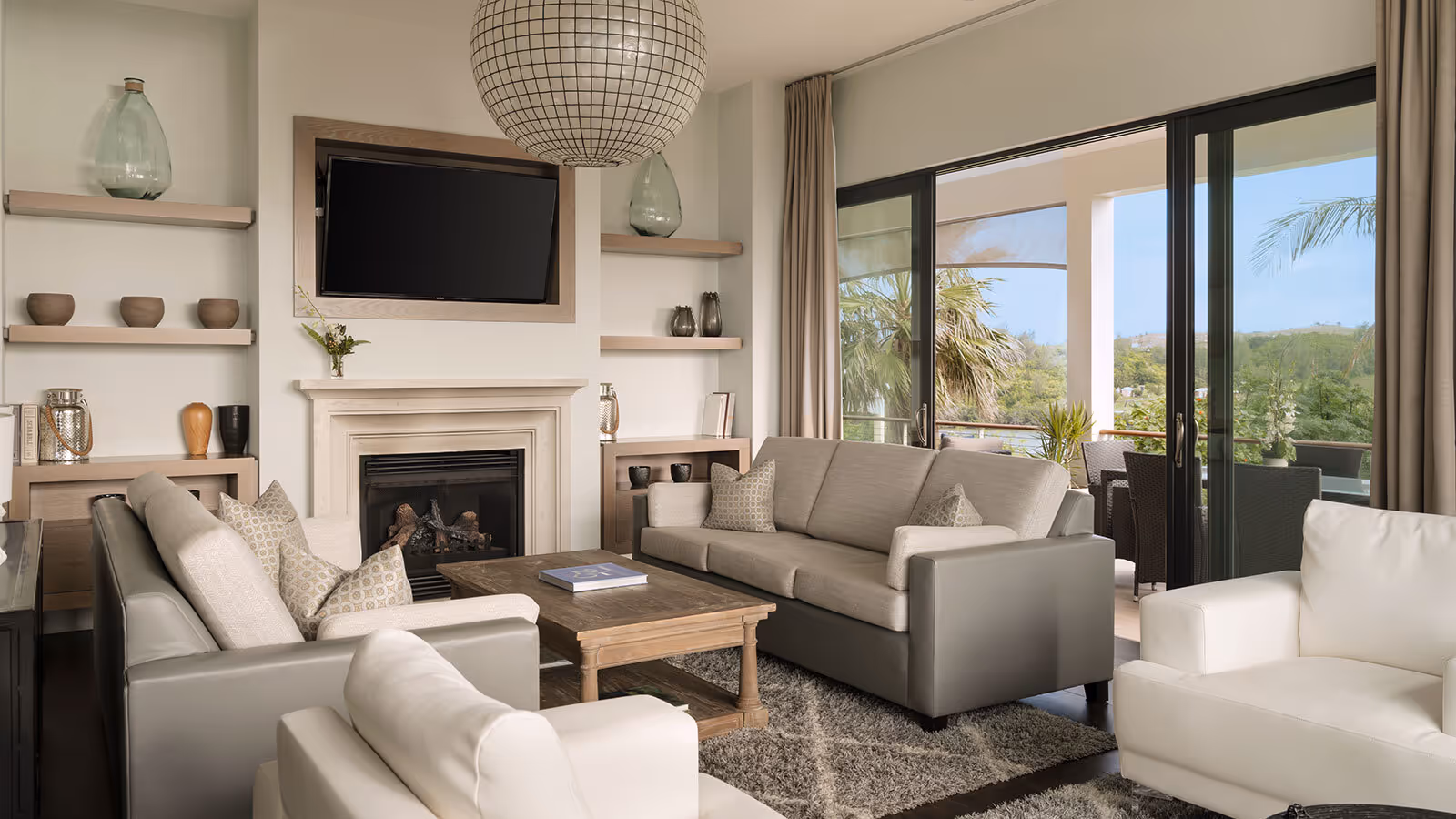 Modern living room with neutral tones, two sofas, armchairs, a fireplace, wall-mounted TV, and large windows to a balcony.