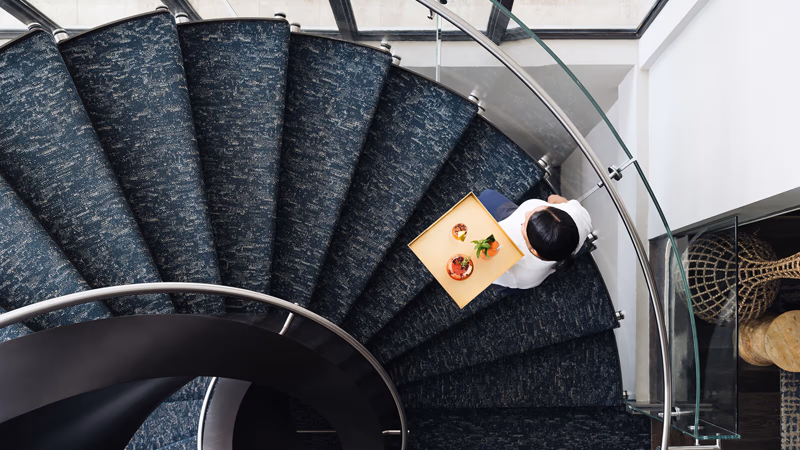 A person carries a tray with food and drinks while walking down a curved, carpeted staircase viewed from above.