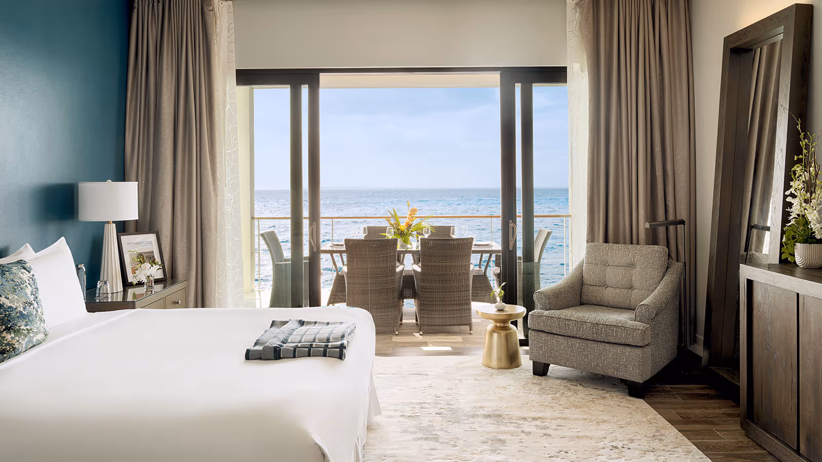 Modern hotel room with a king bed, armchair, and balcony overlooking the ocean with patio seating.