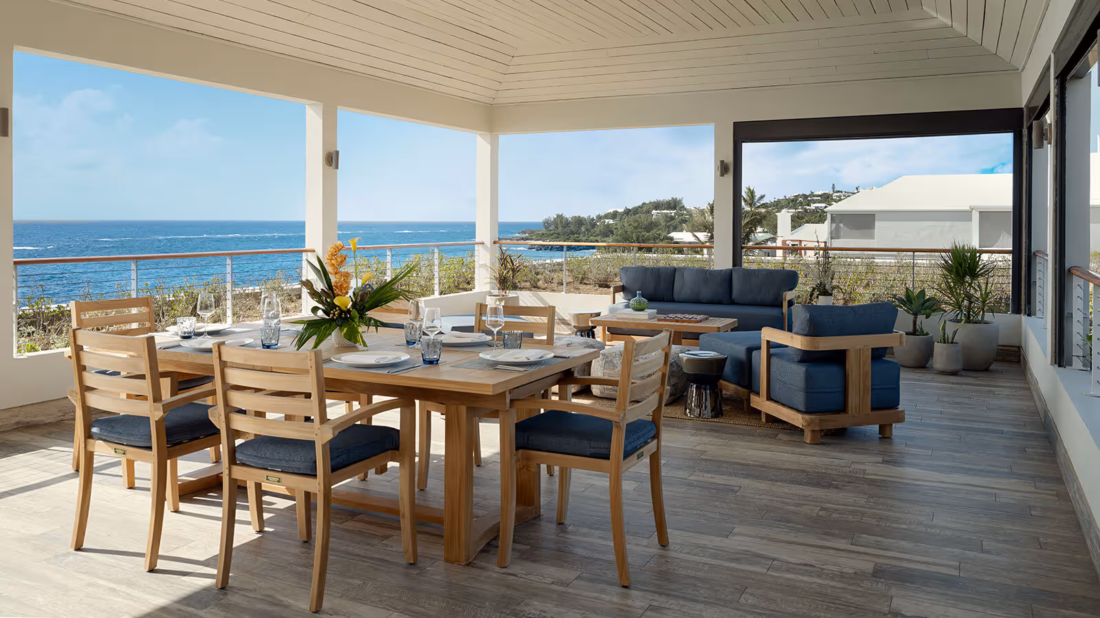 Spacious covered patio with wooden dining set, lounge seating, and ocean view in the background on a sunny day.