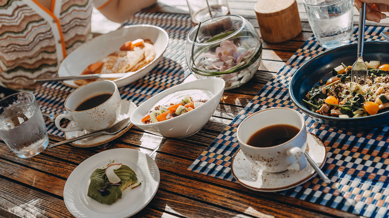 A wooden table set with coffee, salad, avocado toast, fruit bowl, pancakes, and water on patterned placemats.