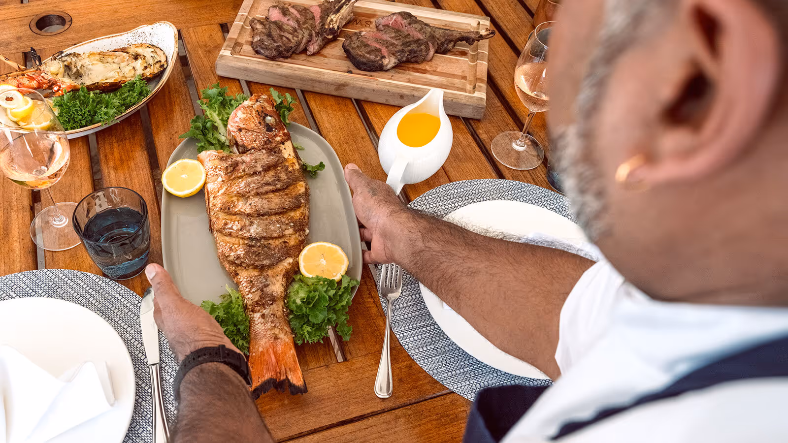 A person places a grilled whole fish with lemon and greens on a dining table set with plates, wine, and other dishes.