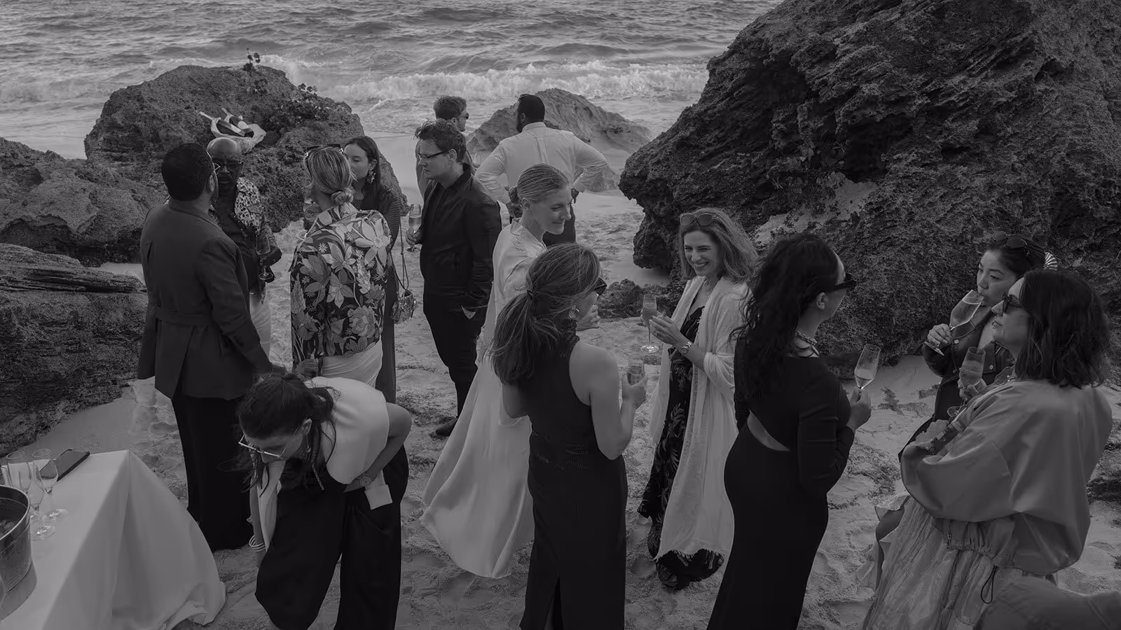 A group of people dressed formally stand and converse by rocky cliffs at a beach, holding drinks near the shoreline.
