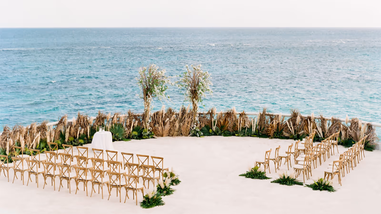 Outdoor wedding ceremony setup by the ocean with chairs arranged in rows and palm decorations in the background.