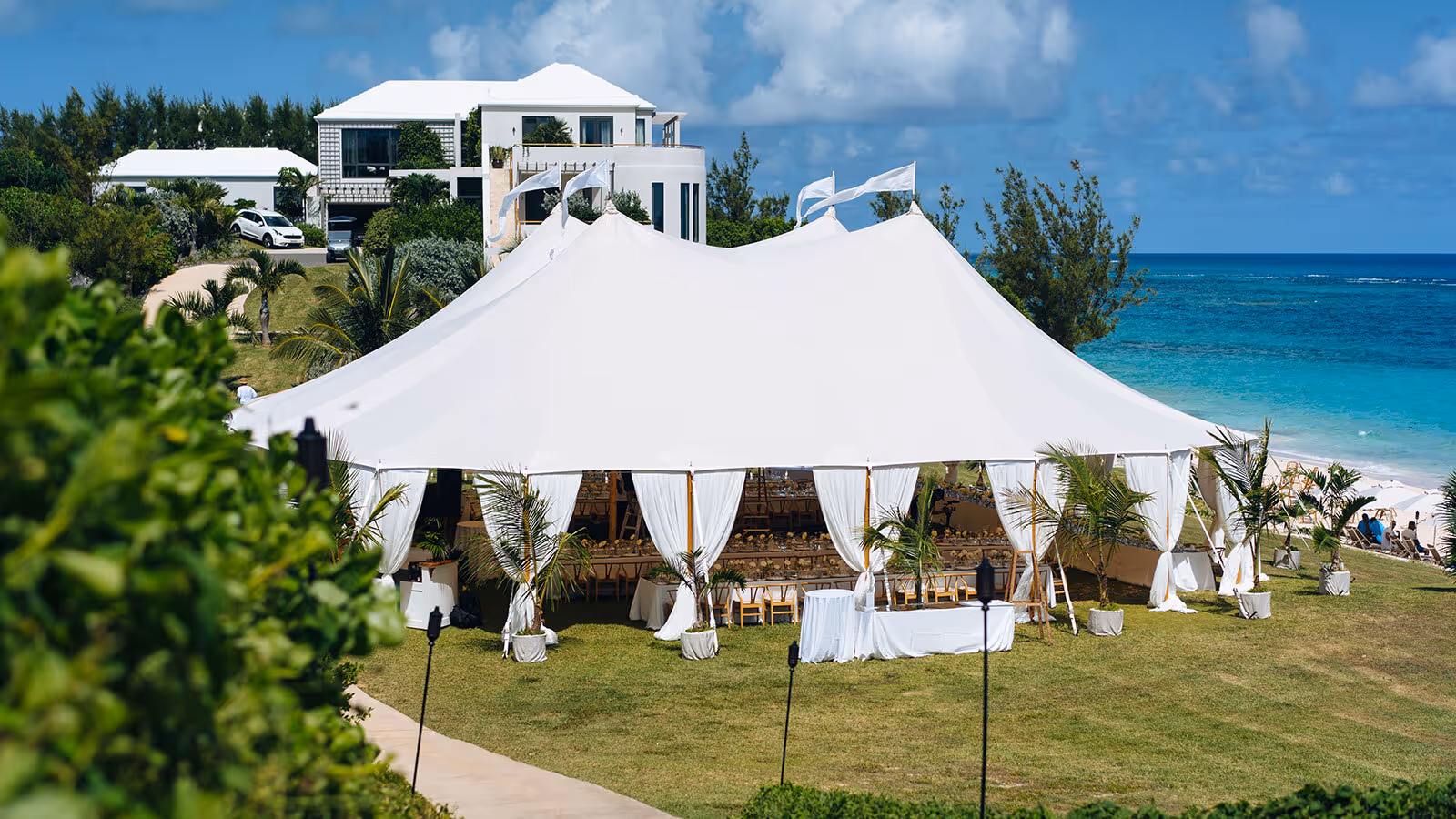 Large white event tent set up on a grassy lawn by the ocean, with a white house and blue sky in the background.