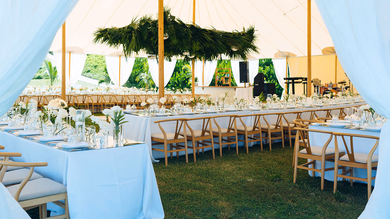 Outdoor event tent with long tables set for dining, white tablecloths, wooden chairs, and green foliage decor overhead.