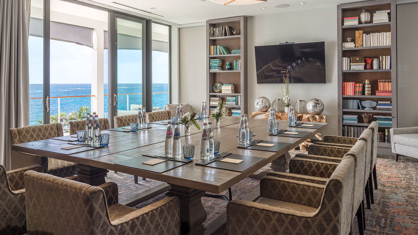 Modern conference room with a large table, chairs, bookshelves, TV, and ocean view through glass doors.