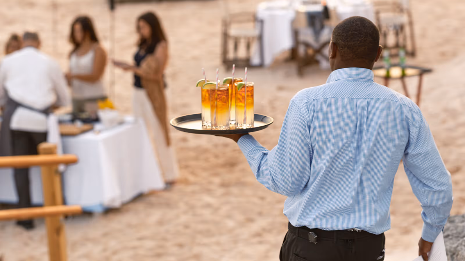 A server carrying a tray of iced drinks approaches a group of people at an outdoor beach event with tables.