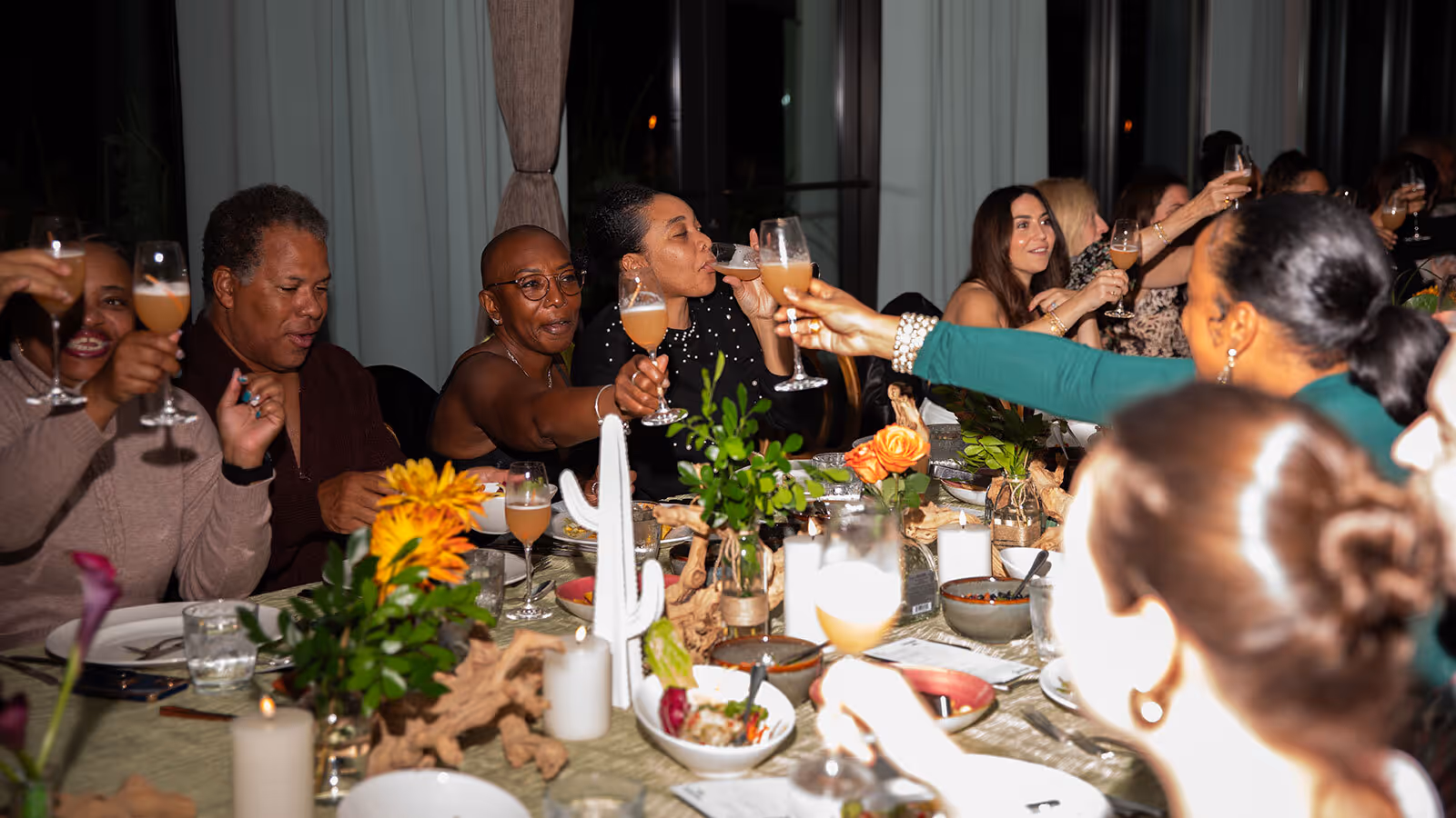 A group of people seated at a dinner table raise glasses in a toast during an indoor gathering.